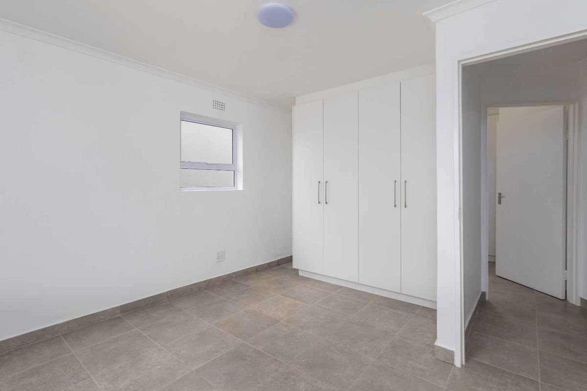 A bright, empty bedroom featuring neutral floor tiles, built-in wardrobes, and a clean, minimalist aesthetic.