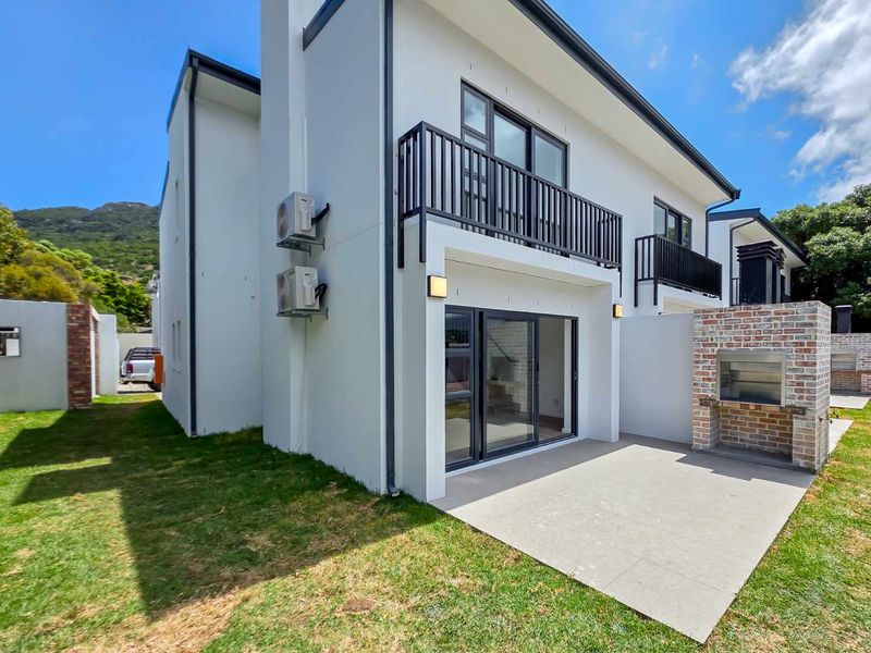 Rear view of a modern townhouse featuring a paved patio with a built-in brick braai, upper-level balcony, and well-maintained lawn area.