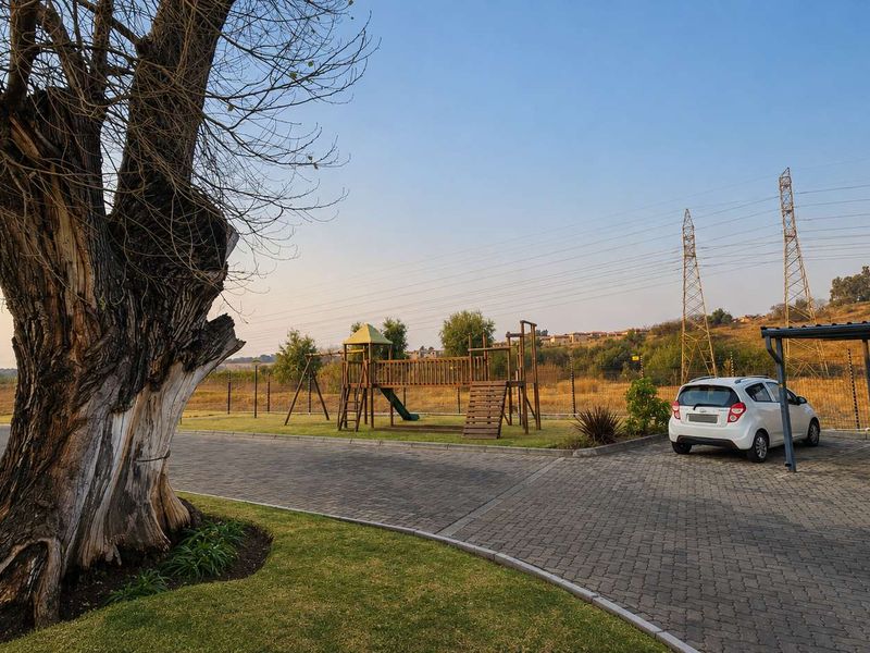 Outdoor communal area featuring a paved driveway, covered carport, and children's playground equipment set within a landscaped garden.