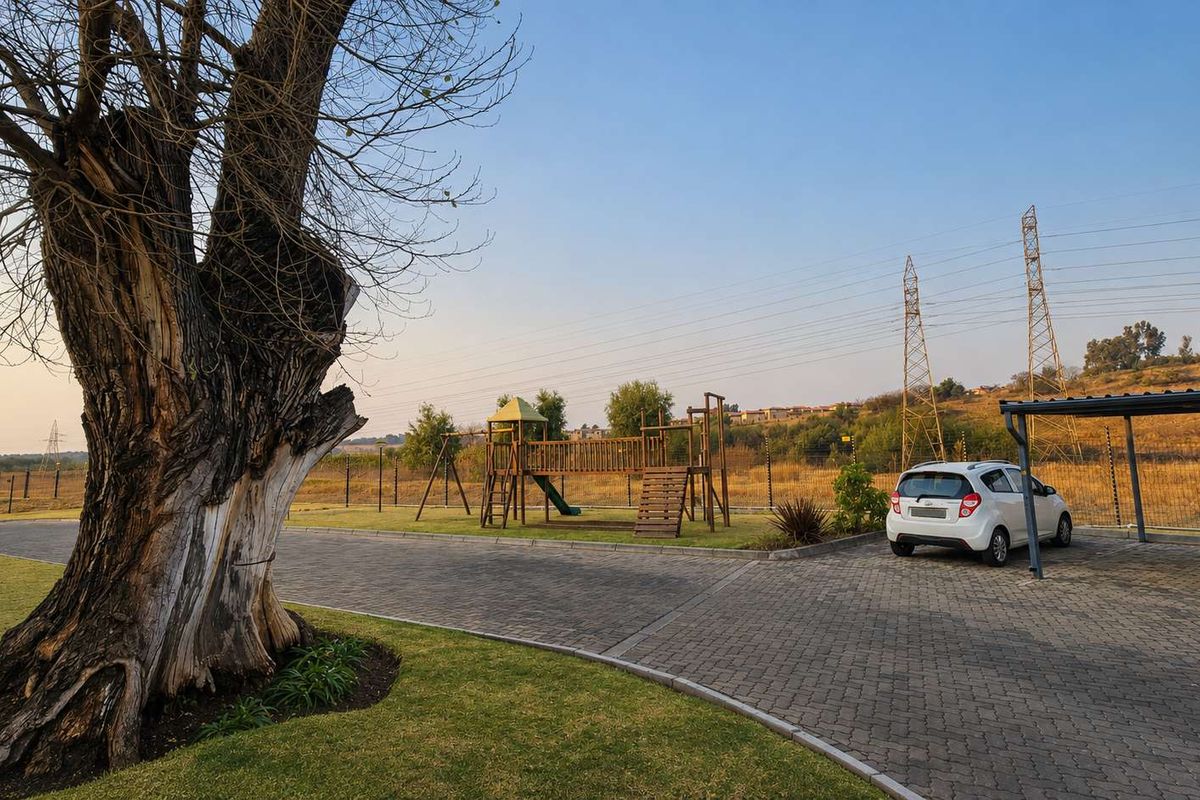 Outdoor communal area featuring a paved driveway, covered carport, and children's playground equipment set within a landscaped garden.