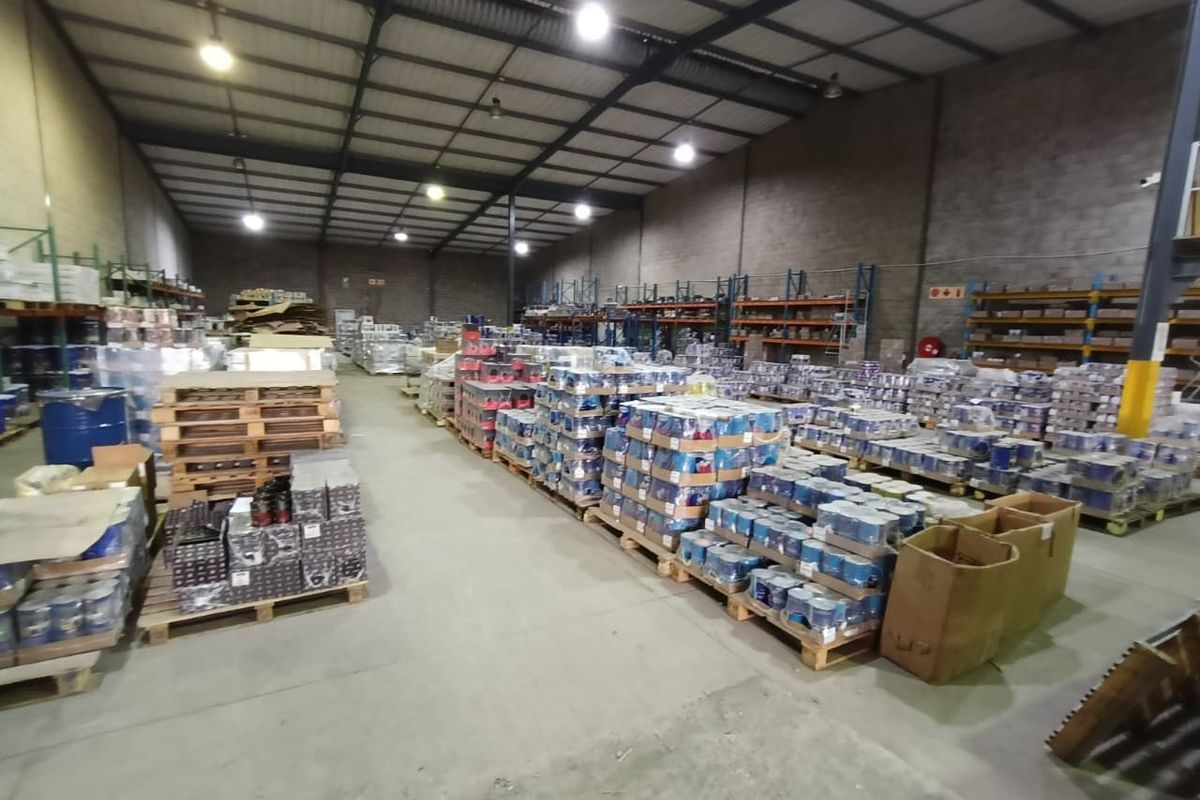 Spacious industrial warehouse featuring high ceilings with exposed steel trusses, concrete flooring, and extensive metal racking systems for storage.