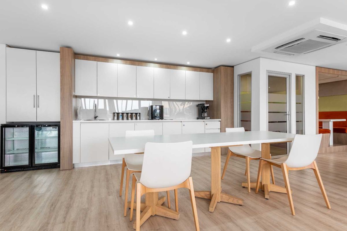 Modern office kitchenette featuring sleek white cabinetry, wood-tone accents, integrated under-counter refrigeration, and recessed ceiling lighting in a clean, professional workspace.