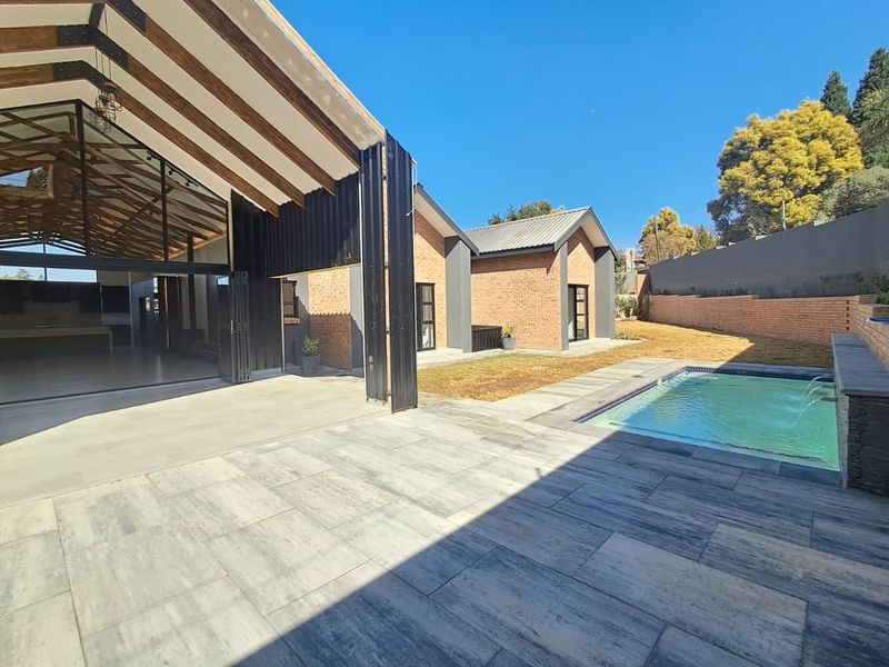 Modern outdoor entertainment area featuring a swimming pool, spacious tiled patio, and a large covered structure with exposed timber beams and brick finishes.