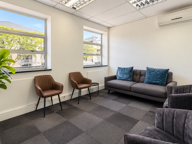 A bright, modern office space featuring carpet tiles, a suspended ceiling with recessed lighting, a split-system air conditioner, and large windows for natural light.