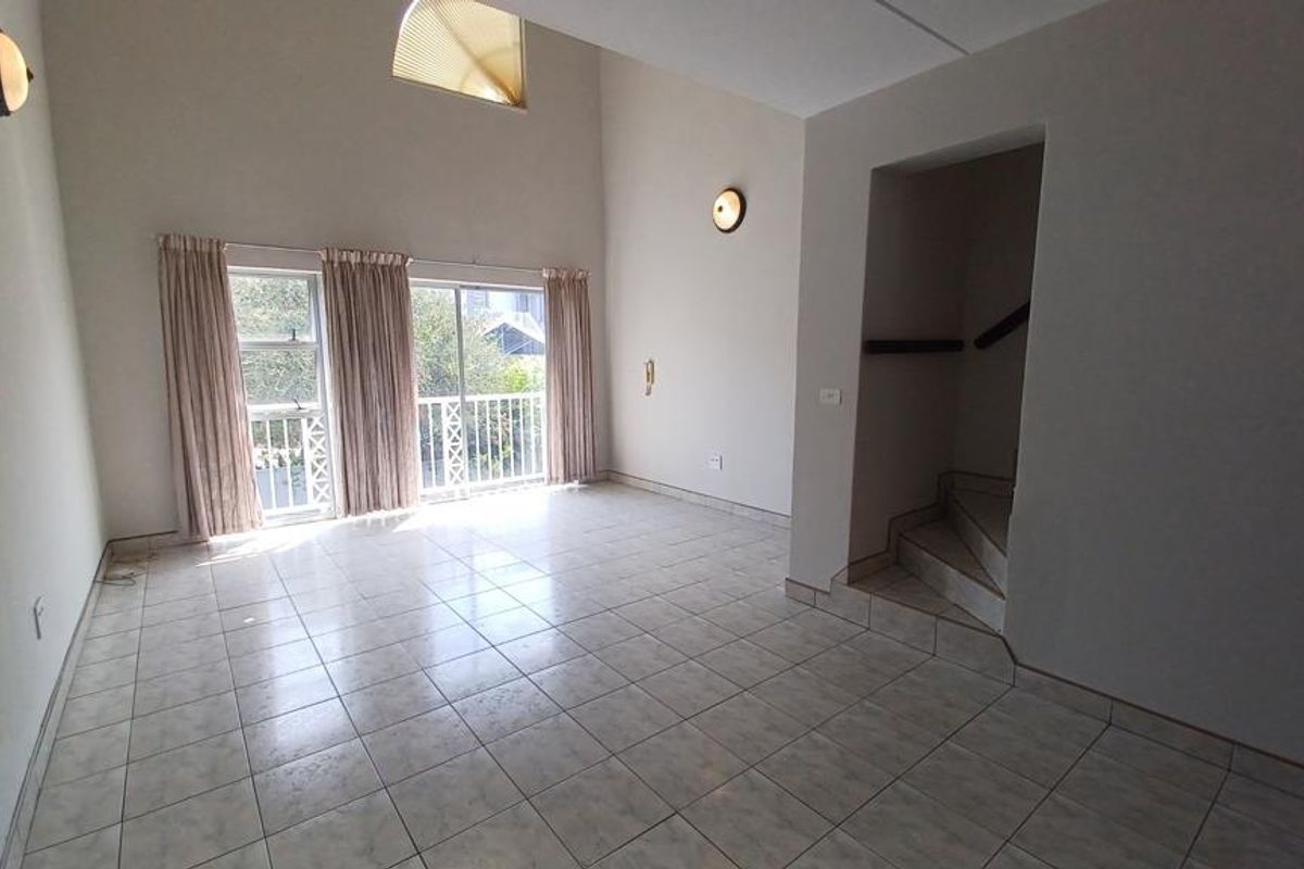 Spacious, light-filled living area featuring high vaulted ceilings, tiled flooring, and sliding glass doors leading to a balcony, with an adjacent staircase.
