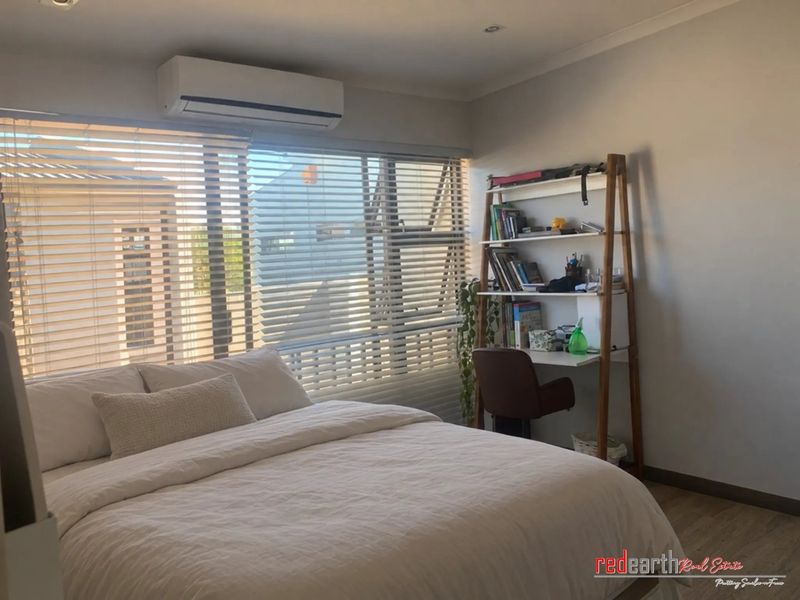 A well-lit bedroom featuring laminate flooring, an integrated air conditioning unit, and large windows fitted with venetian blinds.