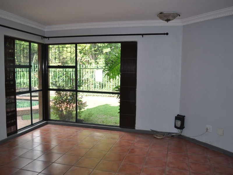 An empty living room featuring terracotta floor tiles, large corner windows with security bars, and a view of the garden.