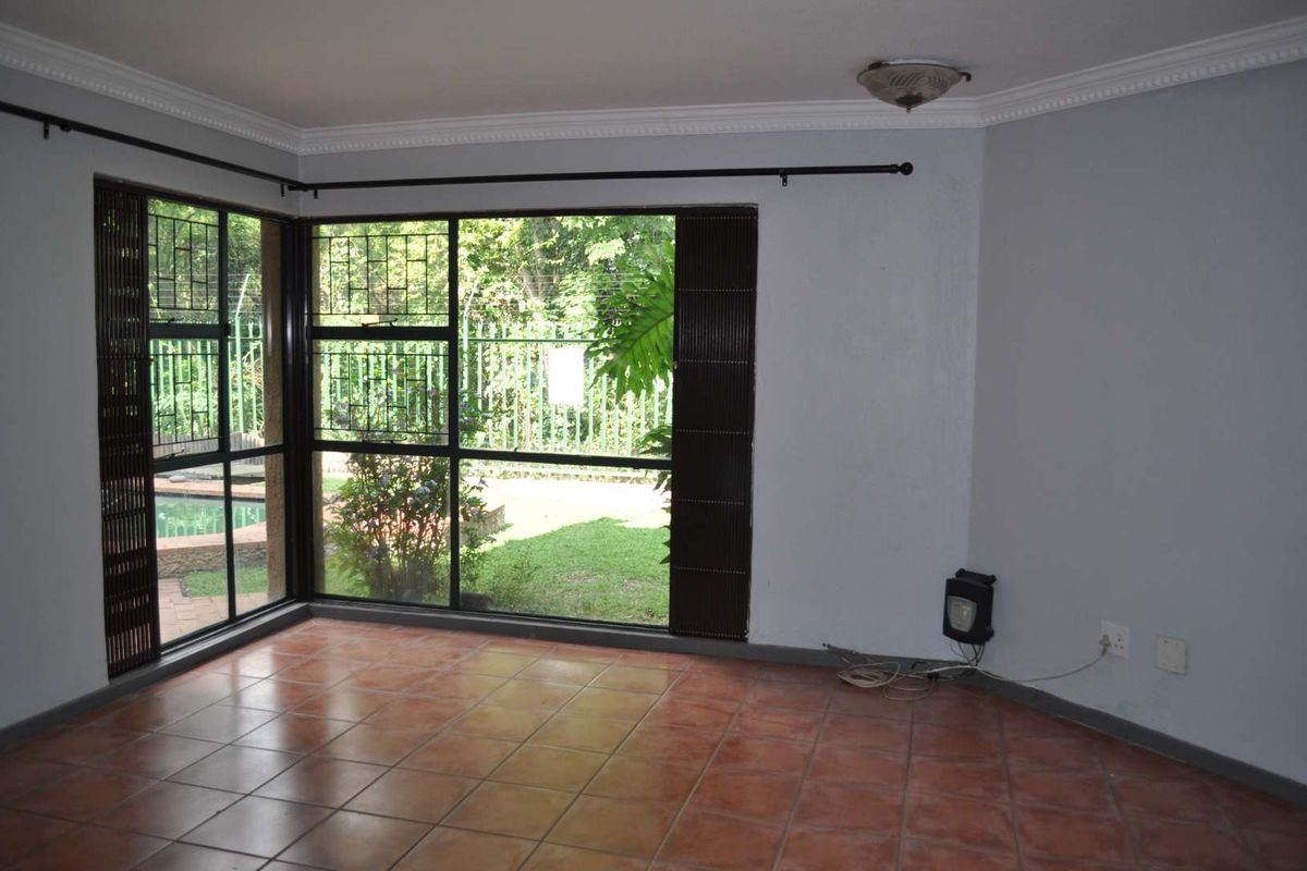 An empty living room featuring terracotta floor tiles, large corner windows with security bars, and a view of the garden.