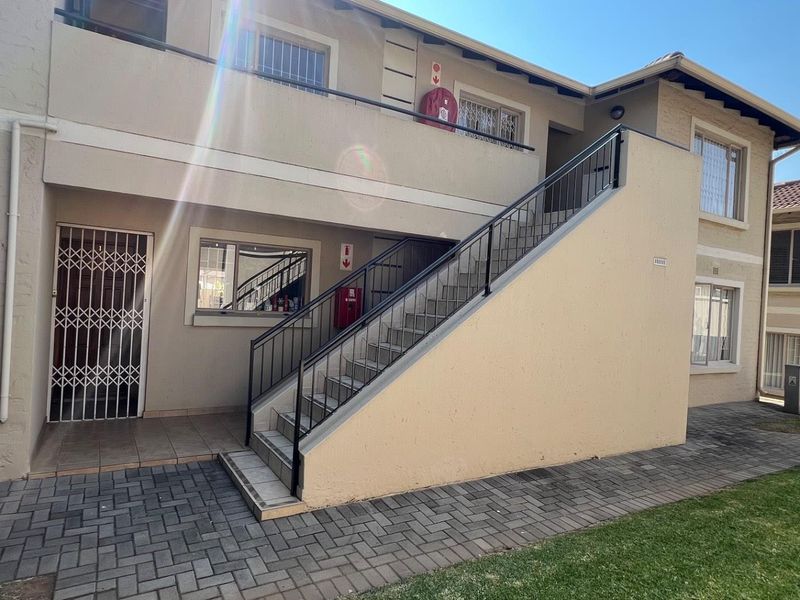 Exterior view of an apartment building featuring a paved walkway, external staircase with metal railing, and security gate access.