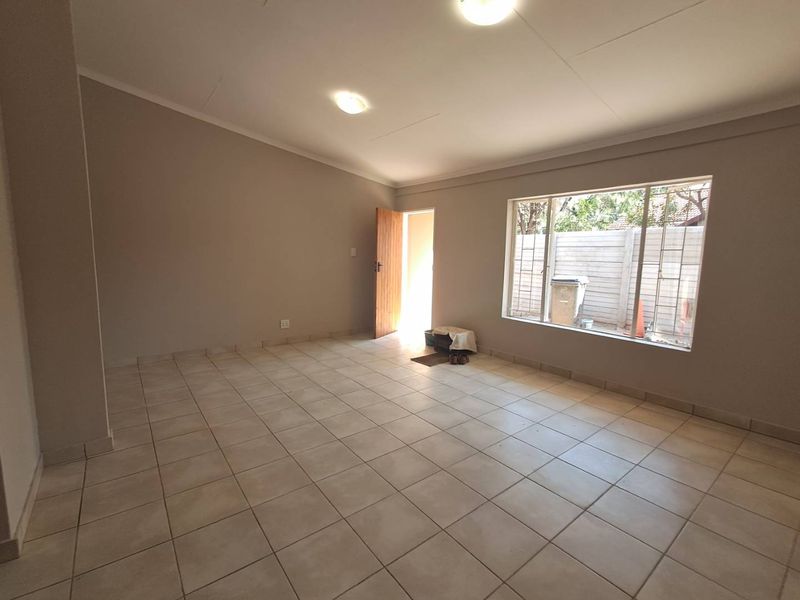 A spacious, empty living area featuring neutral wall tones, tiled flooring, and natural light from a large window with security bars.