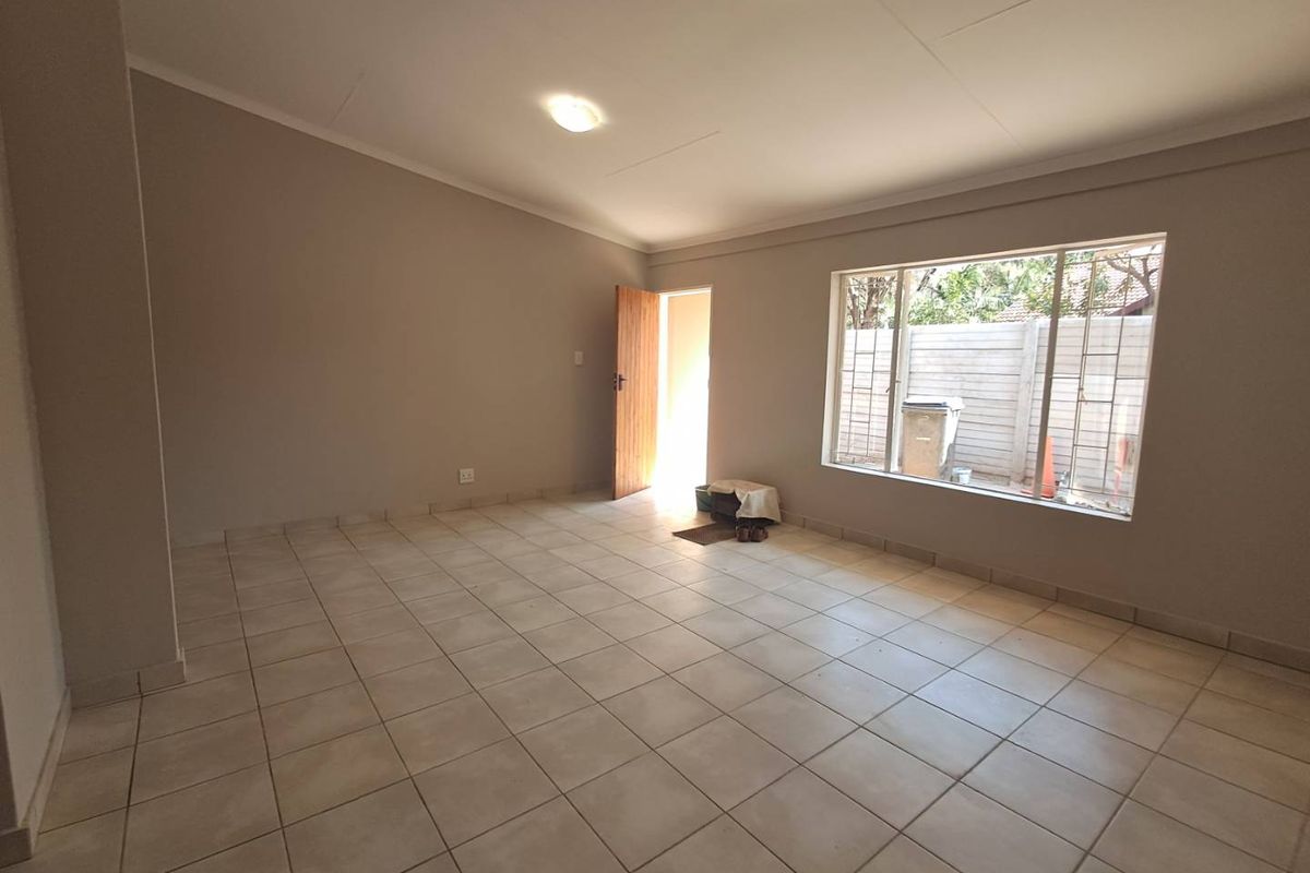 A spacious, empty living area featuring neutral wall tones, tiled flooring, and natural light from a large window with security bars.