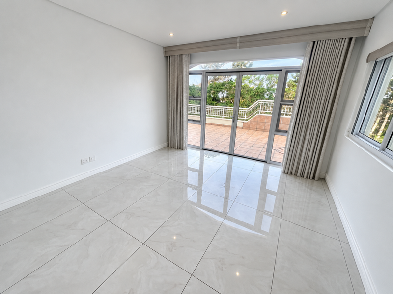 A bright, empty living room featuring large format tiled flooring, recessed lighting, and sliding glass doors leading to an outdoor balcony area.
