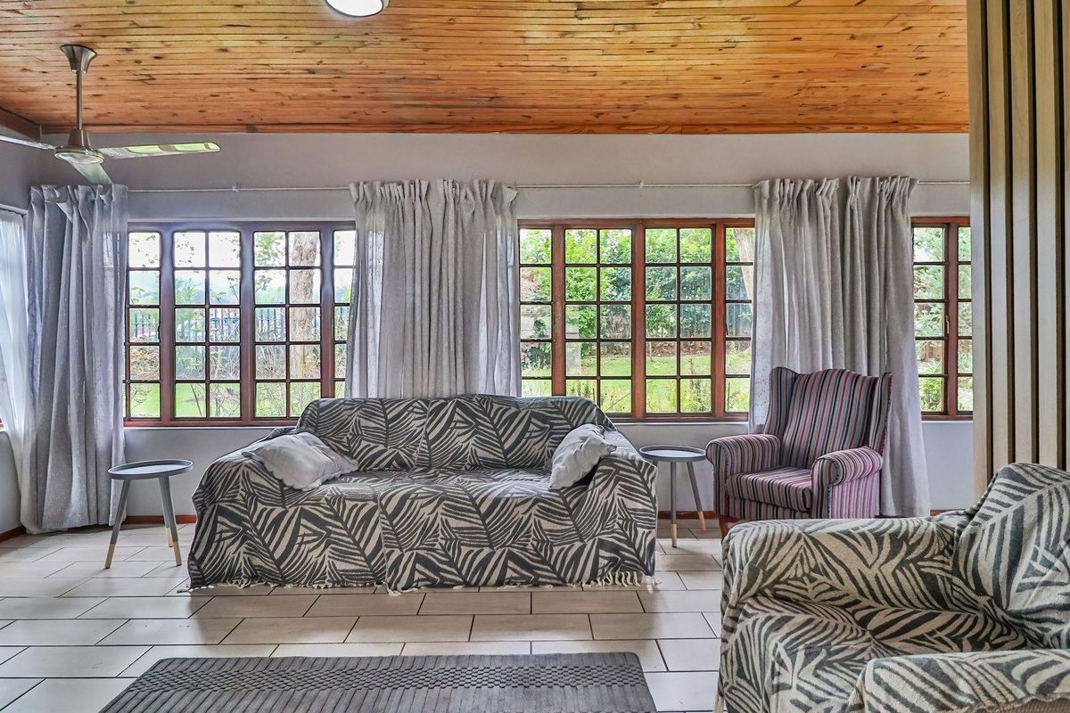 A spacious living room featuring a warm timber-clad ceiling, tiled flooring, and large timber-framed windows that provide ample natural light and garden views.
