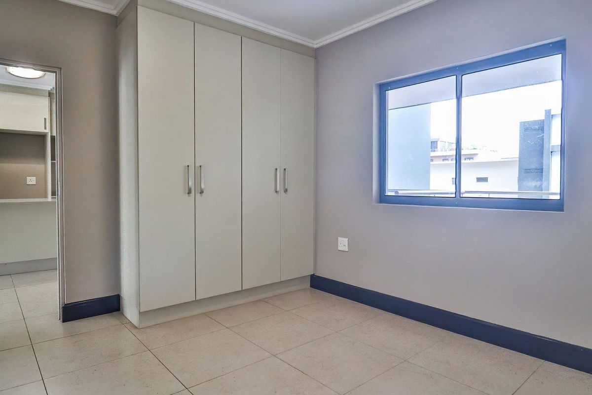 A bright, empty bedroom featuring neutral tiled flooring, built-in cupboards, and a window providing natural light, with a view into an adjacent room.