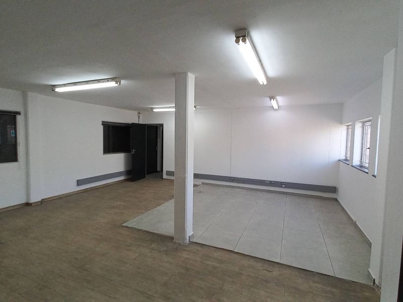 An open-plan industrial office space featuring fluorescent lighting, tiled and vinyl flooring, and multiple windows with security bars.