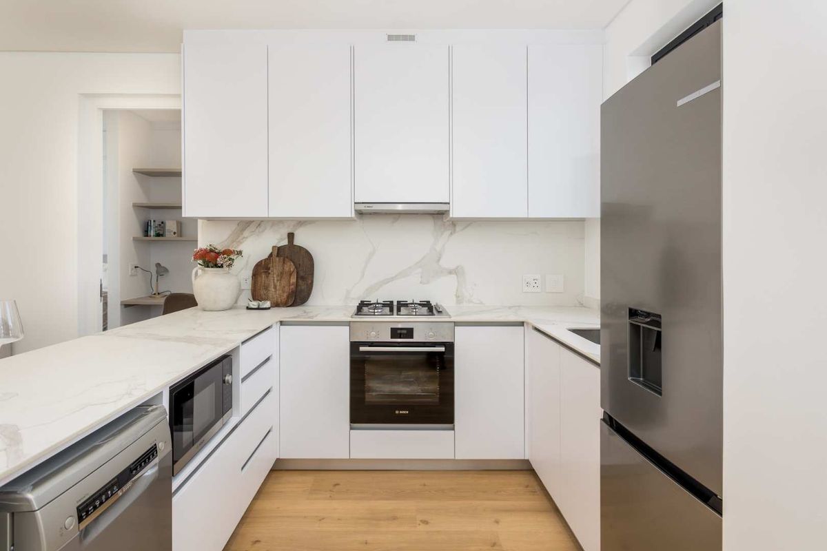 Modern kitchen featuring sleek handleless white cabinetry, marble-look stone countertops and backsplash, integrated appliances, and high-quality engineered wood flooring.