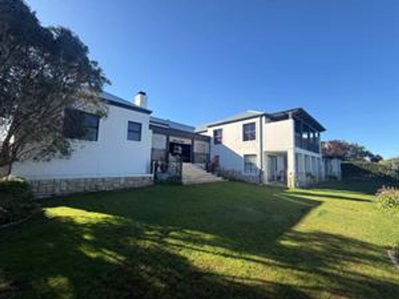 Rear view of a multi-level house featuring white exterior walls, stone cladding, a balcony, and a well-maintained lawn.
