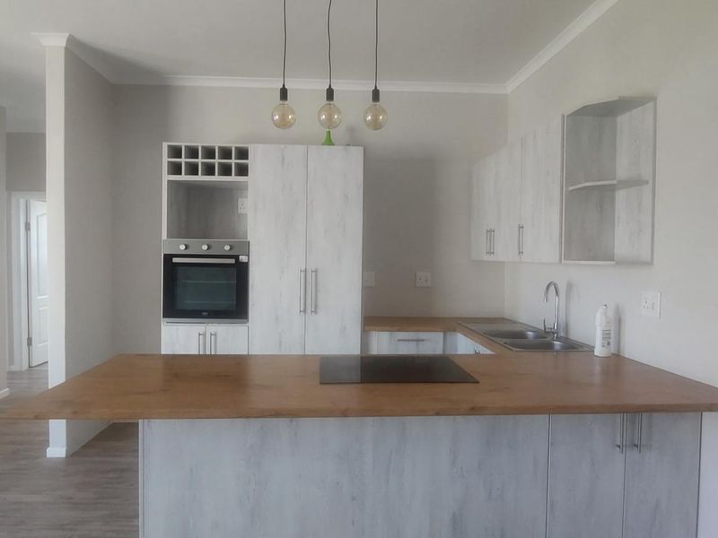 Modern open-plan kitchen featuring light wood-grain cabinetry, a central island with a hob, built-in oven, and double sink, finished with contemporary pendant lighting.