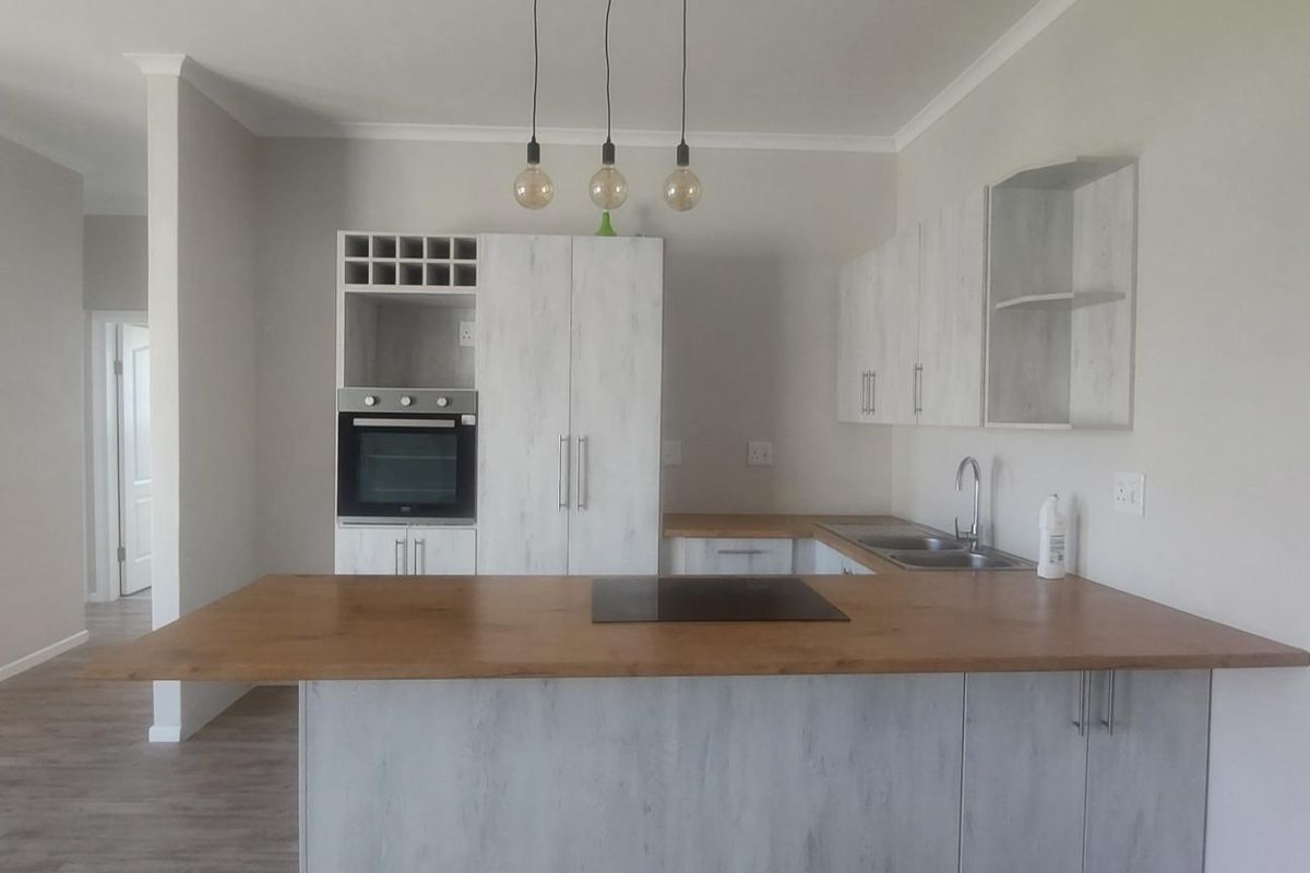 Modern open-plan kitchen featuring light wood-grain cabinetry, a central island with a hob, built-in oven, and double sink, finished with contemporary pendant lighting.