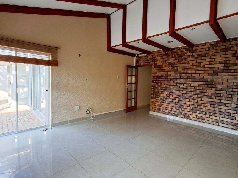Spacious living area featuring exposed timber ceiling beams, a rustic brick feature wall, tiled flooring, and direct access to the outdoor patio.