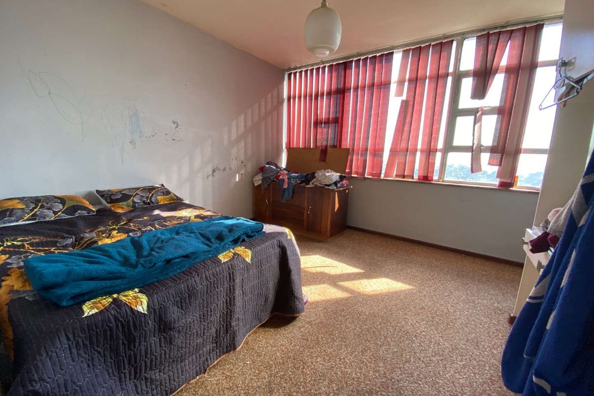 A bedroom featuring large windows with natural light, carpeted flooring, and a pendant light fixture, currently requiring cosmetic updates and wall repairs.