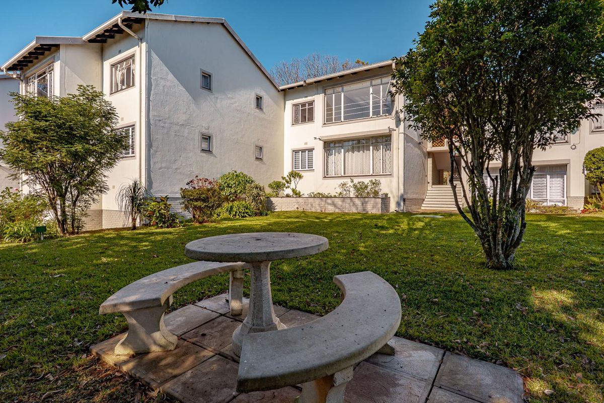 A view of the rear exterior of the apartment building, featuring a landscaped garden area with a fixed concrete table and bench set.