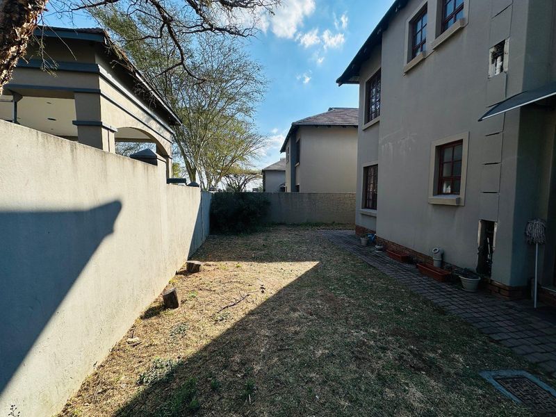 A rear exterior view of the townhouse featuring a paved walkway, boundary wall, and a small garden area with established trees.