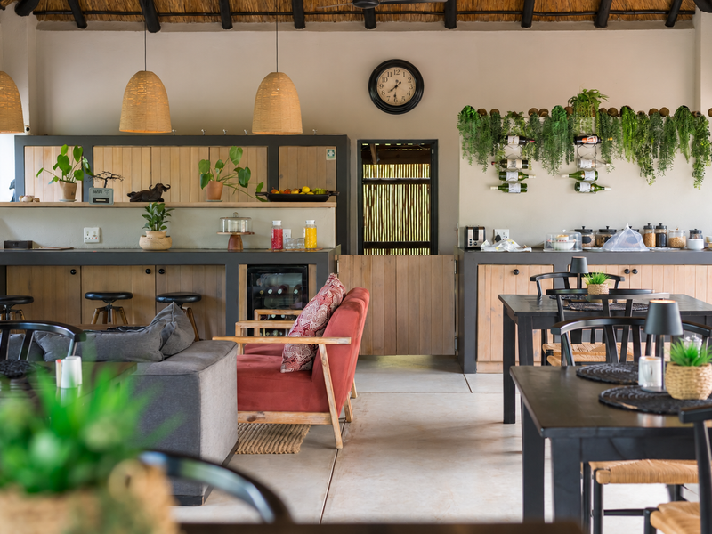 An open-plan guest house dining and lounge area featuring a rustic thatched roof, exposed timber rafters, and custom built-in cabinetry with a stable door.
