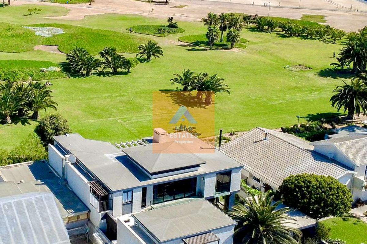 Aerial view of a modern multi-level coastal property featuring flat roofs, balconies, and proximity to the beach and parkland.