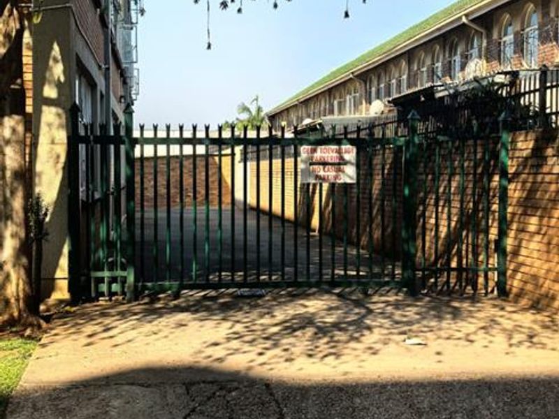 Exterior view of the apartment complex entrance featuring a secure metal gate, brick boundary wall, and concrete driveway access.