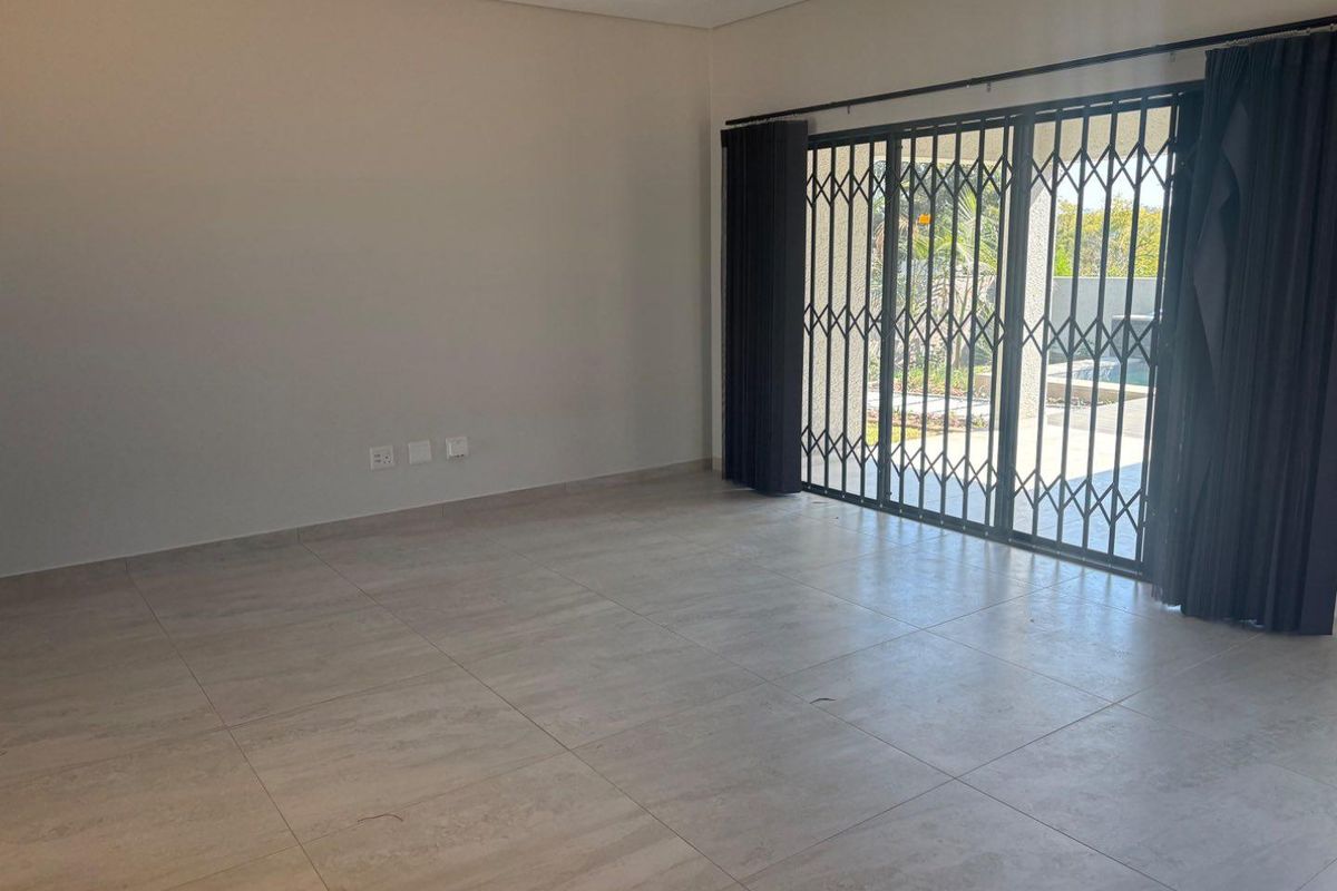 Spacious, empty living area featuring neutral tiled flooring and a sliding glass door with a security gate leading to the outdoor patio.