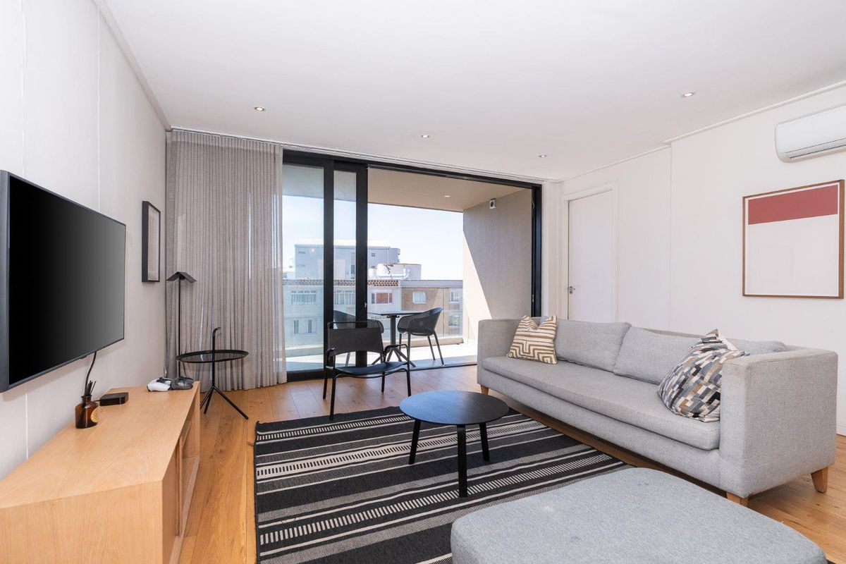 Modern living room featuring timber flooring, recessed downlighting, wall-mounted air conditioning unit, and large sliding glass doors opening onto a private balcony.
