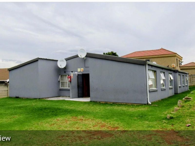 Front exterior view of a single-story townhouse featuring a grey rendered facade, security gate, and surrounding lawn area.