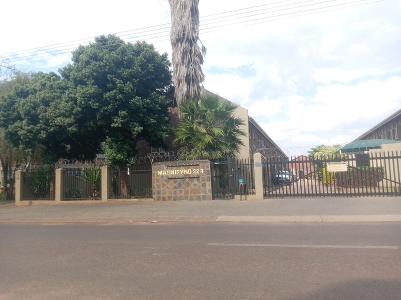 Street view of the apartment complex entrance featuring a stone-clad pillar, security fencing with razor wire, and a gated driveway access point.