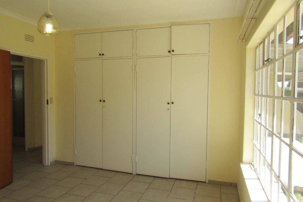 A bright, empty bedroom featuring tiled flooring, large steel-framed windows, and extensive built-in cupboard storage.
