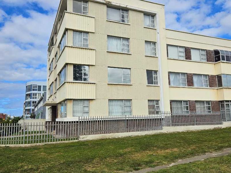 Exterior view of a multi-story apartment building featuring a cream-colored facade, steel-framed windows, and perimeter security fencing.