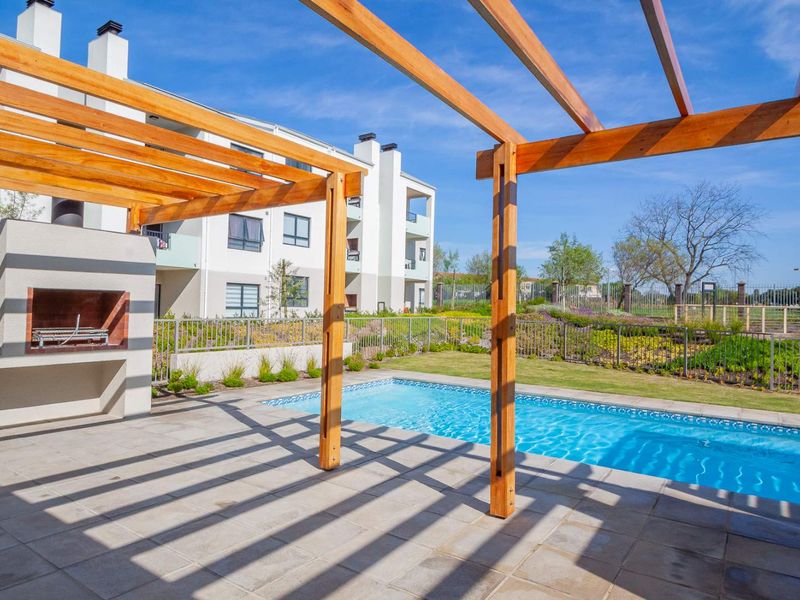 Outdoor pool area featuring a paved patio, wooden pergola, and a built-in braai, set within a well-maintained apartment complex garden.