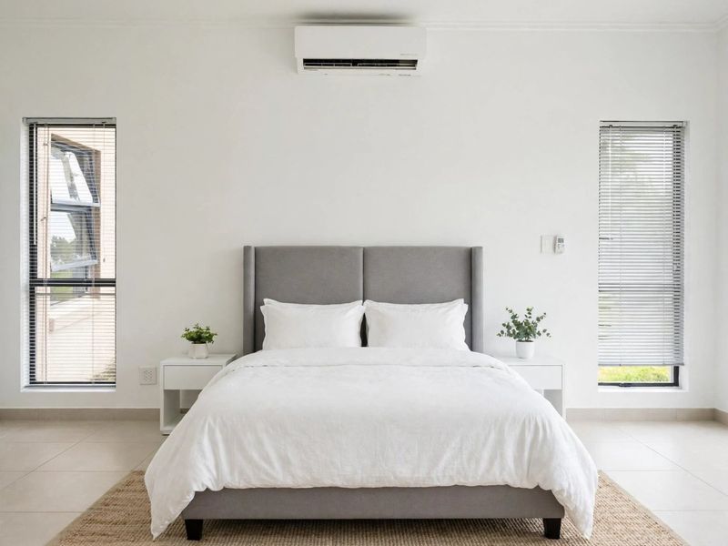 A bright and modern bedroom featuring tiled flooring, dual casement windows with venetian blinds, and a wall-mounted split-system air conditioner.