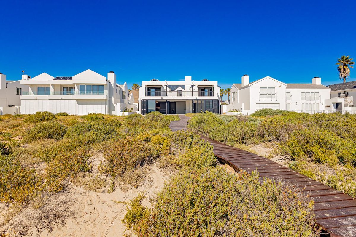 Rear view of the property featuring a wooden boardwalk leading through coastal vegetation to the modern multi-story residence with balconies and sliding doors.