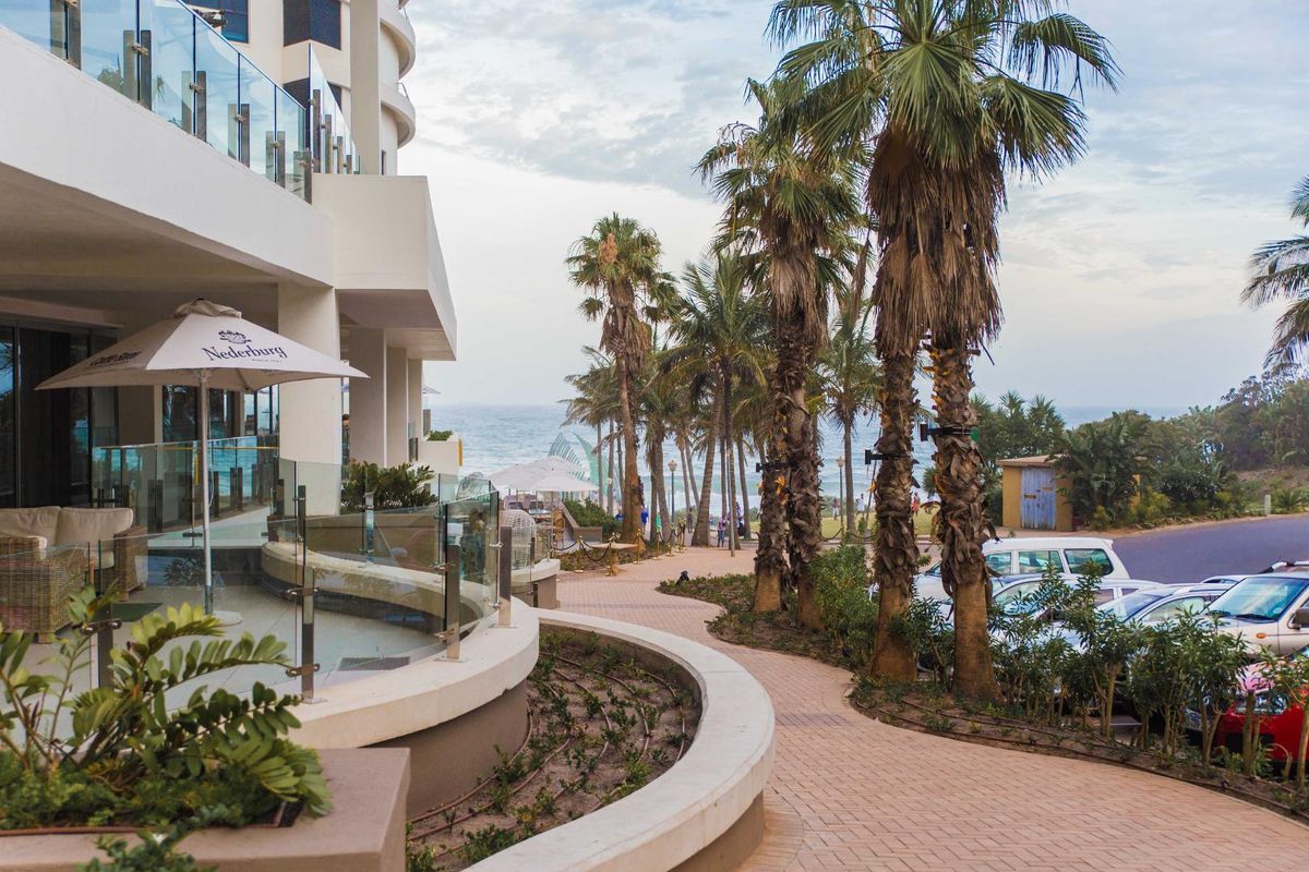 A view of the apartment building exterior featuring a paved walkway, landscaped palm trees, and a glass-railed patio area overlooking the ocean.