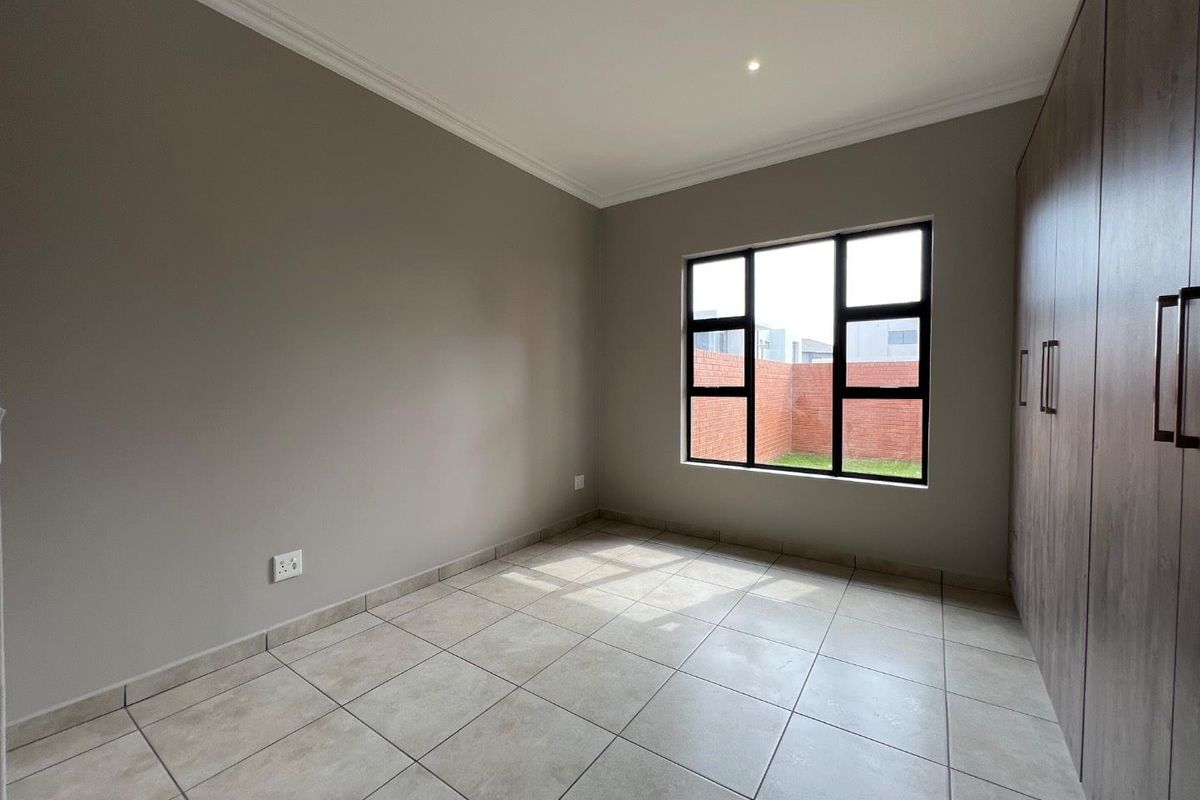 A bright, empty bedroom featuring neutral tiled flooring, built-in cupboards, and large windows overlooking a private rear garden area.