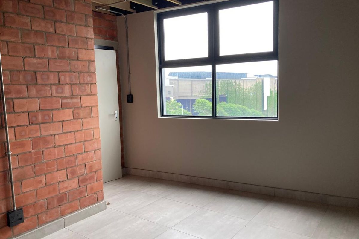 An empty bedroom featuring industrial-style exposed brickwork, tiled flooring, and an exposed ceiling structure with a large window providing natural light.