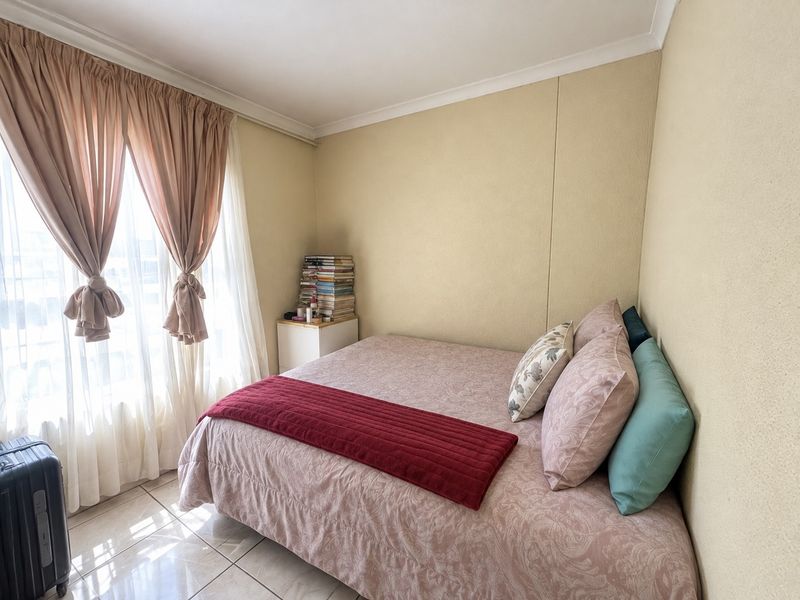 A compact bedroom featuring tiled flooring and neutral wall finishes, illuminated by natural light through a large window with curtain treatments.