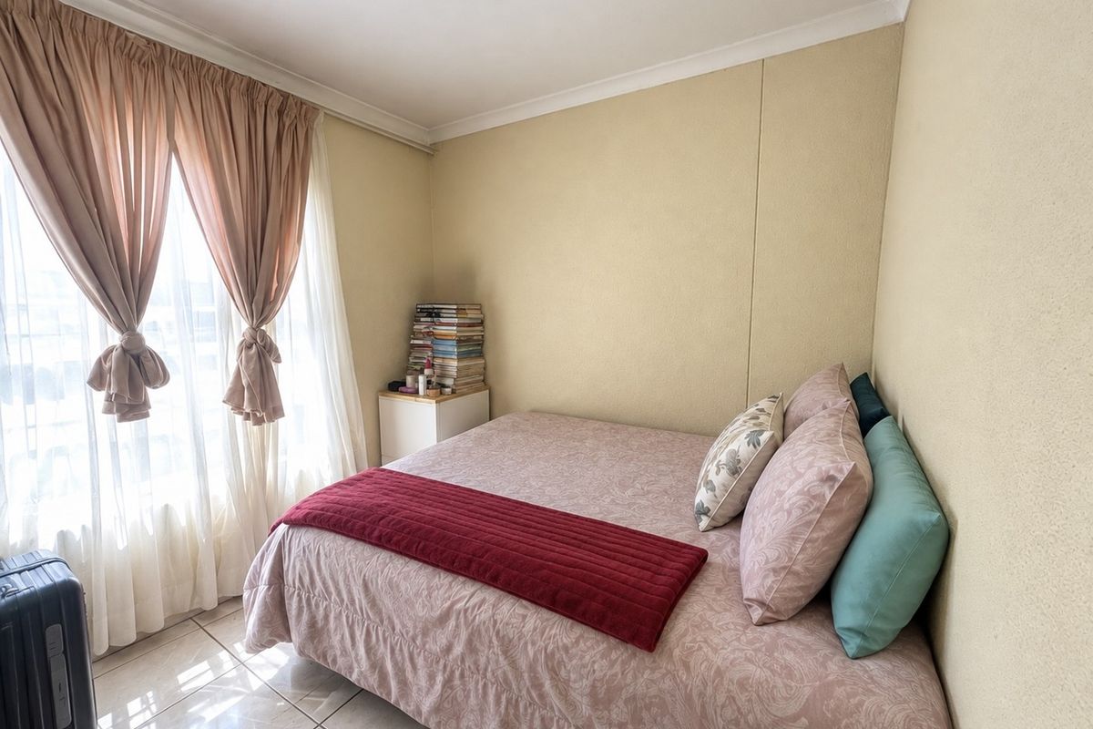 A compact bedroom featuring tiled flooring and neutral wall finishes, illuminated by natural light through a large window with curtain treatments.