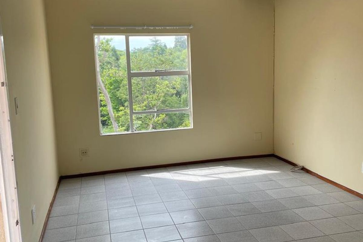 A bright, empty bedroom featuring neutral wall tones, tiled flooring, and a large window providing natural light and a view of the surrounding greenery.