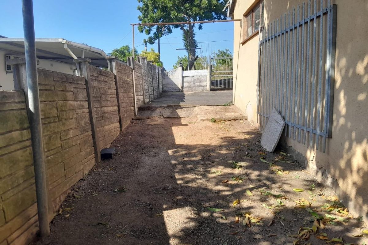 A narrow side or rear exterior passage featuring a precast concrete boundary wall, metal security fencing, and a concrete driveway area requiring maintenance.