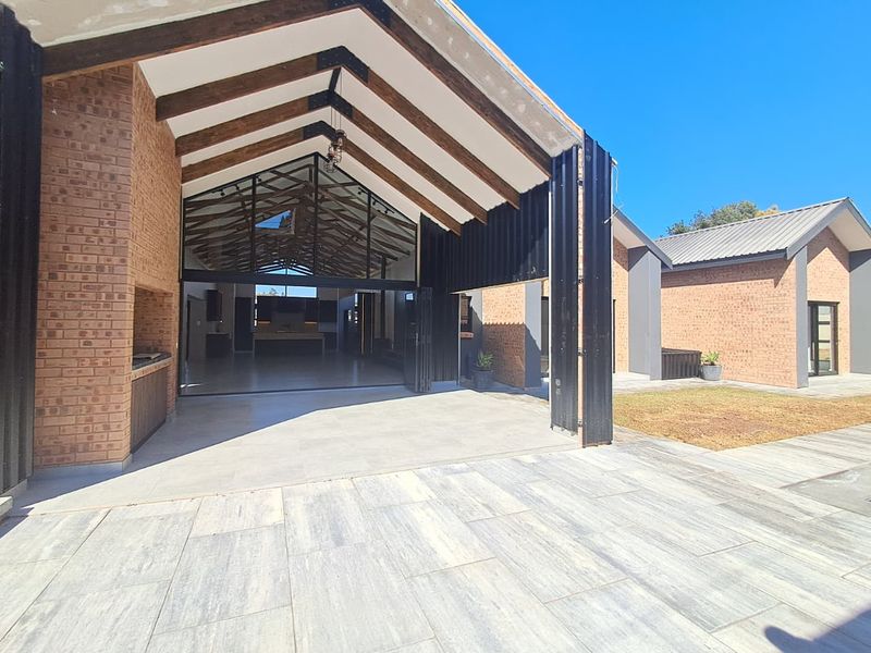 Spacious covered patio featuring exposed timber trusses, built-in braai, and seamless flow through folding glass doors into the main living area.