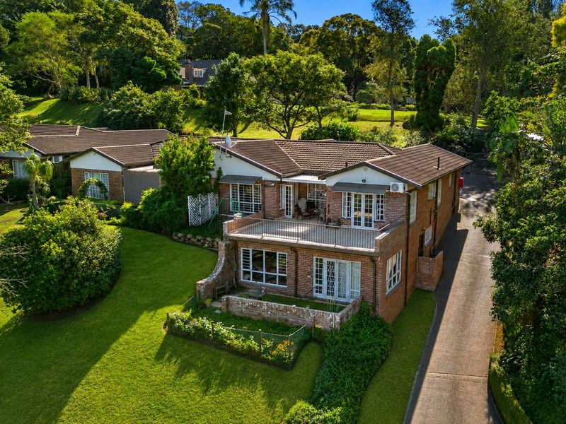 Aerial view of a brick townhouse featuring a tiled roof, upper-level balcony, ground-floor patio, and surrounding landscaped garden area.