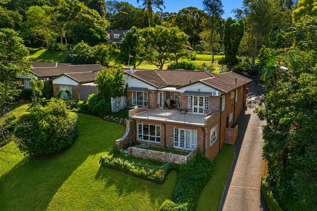 Aerial view of a brick townhouse featuring a tiled roof, upper-level balcony, ground-floor patio, and surrounding landscaped garden area.