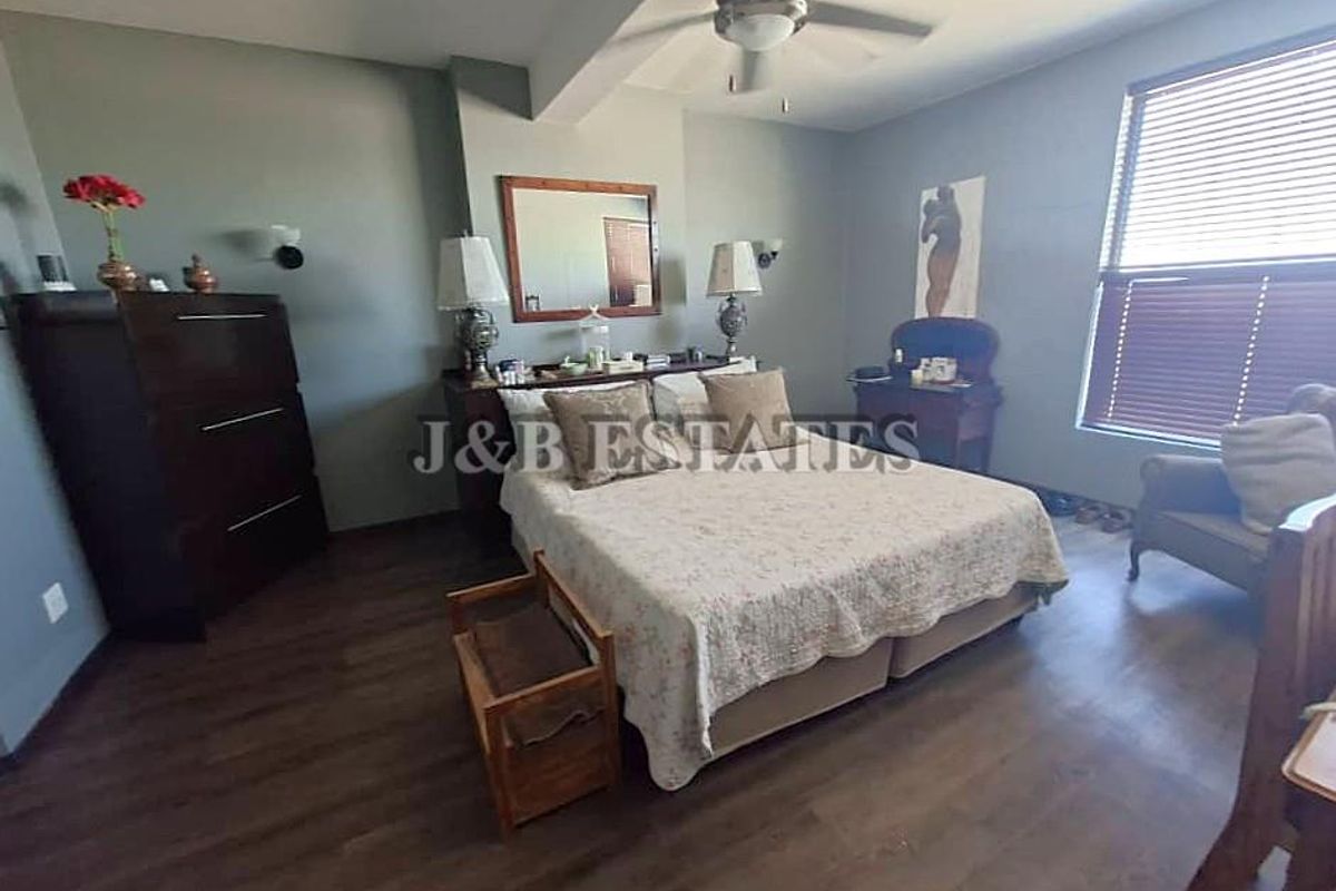 A spacious bedroom featuring dark wood-look flooring, a ceiling fan, and large windows with fitted blinds.